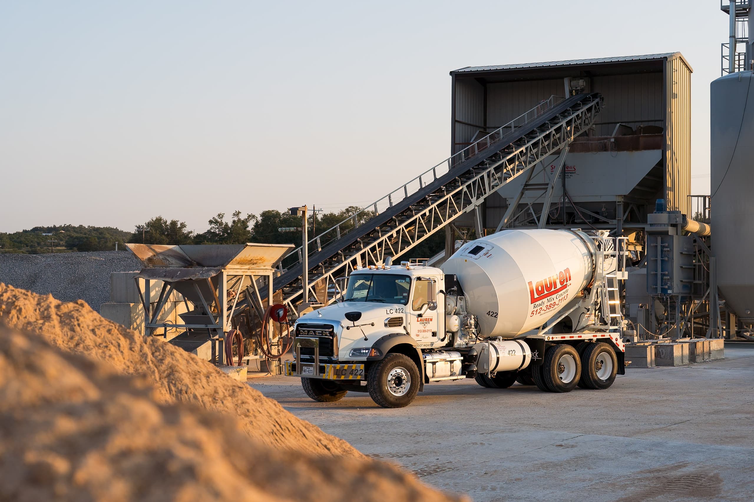 A Lauren Concrete Ready-Mix Truck in Front of the Conveyer Belt at the Plant with a Pile Sand in the Forefront of the Photo.